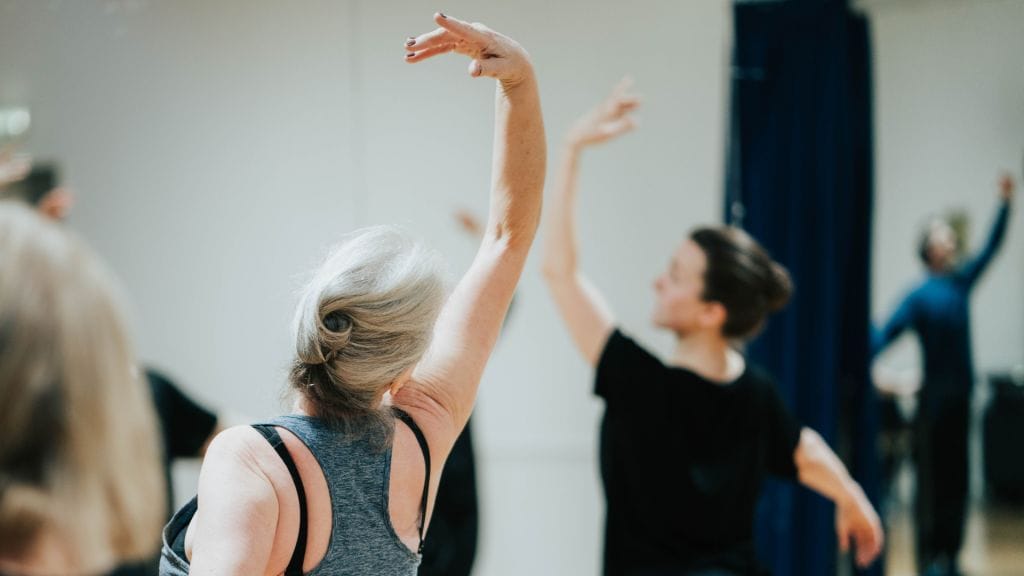 A person stands with their back to the camera, their arm is stretched elegantly above their head in a ballet pose. They are looking towards a ballet instructor, who is out of focus in the background but is doing the same pose.