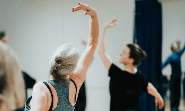 A person stands with their back to the camera, their arm is stretched elegantly above their head in a ballet pose. They are looking towards a ballet instructor, who is out of focus in the background but is doing the same pose.