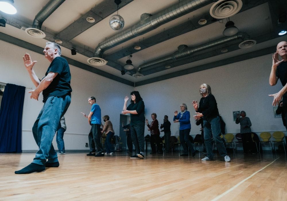Several people in a Tai Chi pose. Their right legs are all outstretched, with their arms raised slightly, all of them are looking intently ahead and focused.