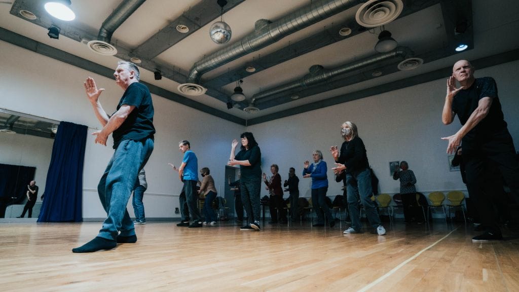 Several people in a Tai Chi pose. Their right legs are all outstretched, with their arms raised slightly, all of them are looking intently ahead and focused.