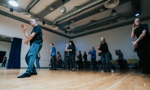 Several people in a Tai Chi pose. Their right legs are all outstretched, with their arms raised slightly, all of them are looking intently ahead and focused.