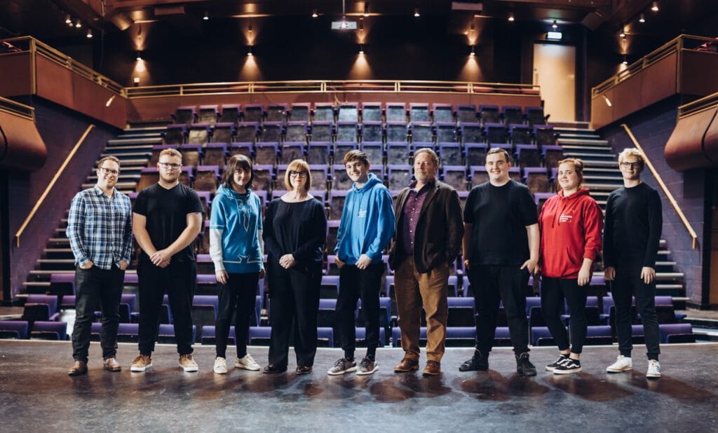 A group of people stand on a stage with rows of empty theatre seats behind them.