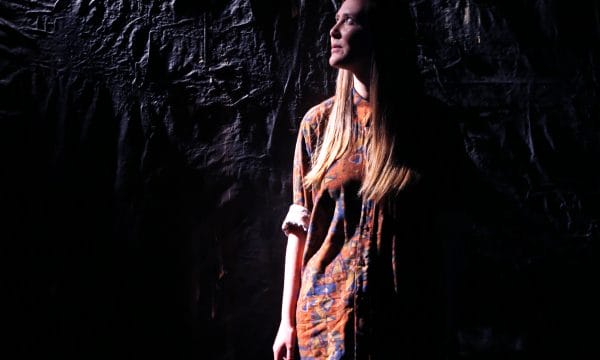 ARC Associate Artist stands in partial darkness, against a black wall. She is looking up to her right, from where a light source casts light on her face and the upper part of her body.