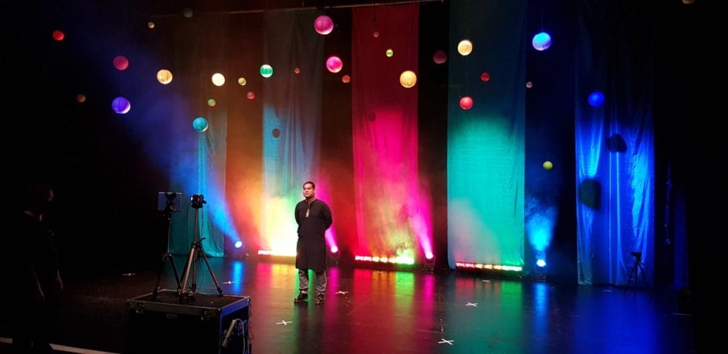 An asian man stands on a stage surrounded by hanging paper lanterns and banners made from colourful cloth.