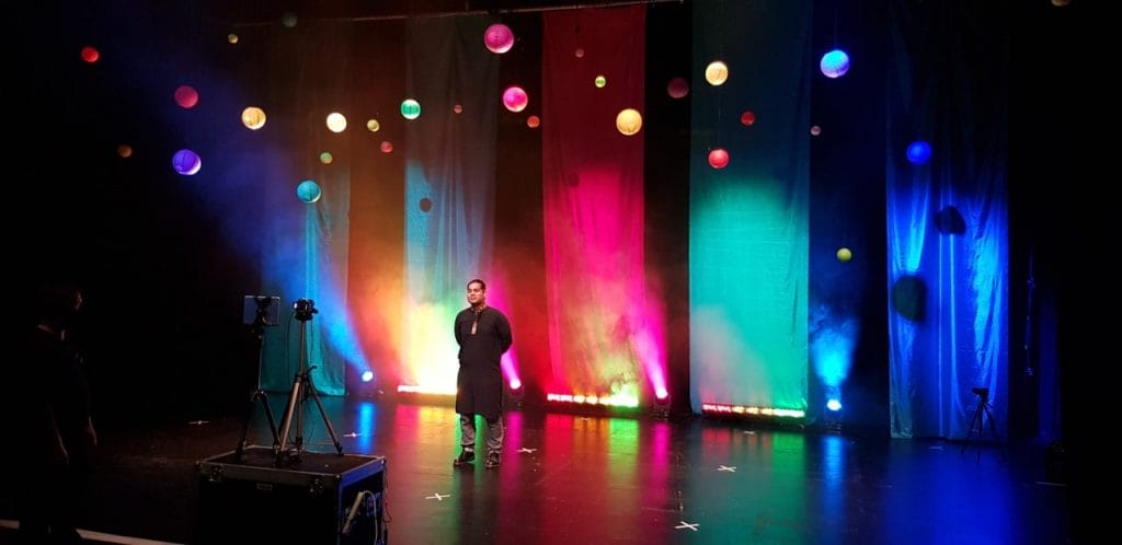 An asian man stands on a stage surrounded by hanging paper lanterns and banners made from colourful cloth.