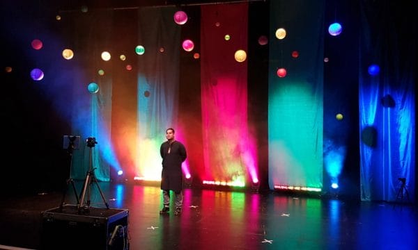 An asian man stands on a stage surrounded by hanging paper lanterns and banners made from colourful cloth.