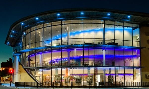 Exterior image of ARC, a building with curved glass frontage over three-floors on the corner of a road intersection. The building is it up at night-time. Logo in the bottom of the image is for EnA Photography.