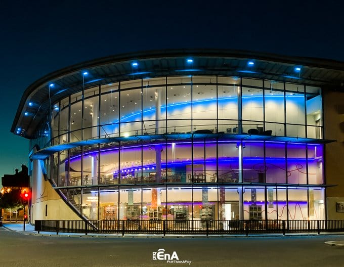 Exterior image of ARC, a building with curved glass frontage over three-floors on the corner of a road intersection. The building is it up at night-time. Logo in the bottom of the image is for EnA Photography.