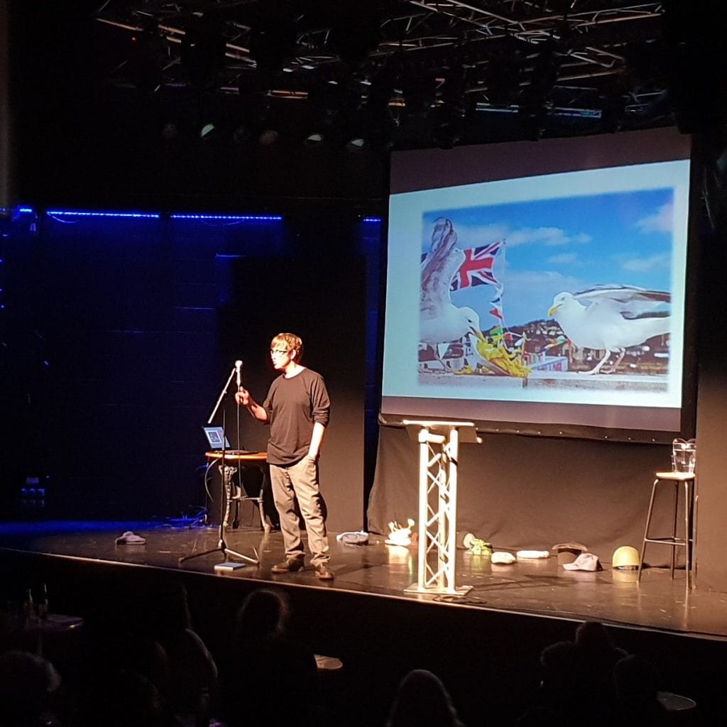 Mike Edwards stands on stage facing an audience. There is a picture of seagulls projected onto a screen behind him.