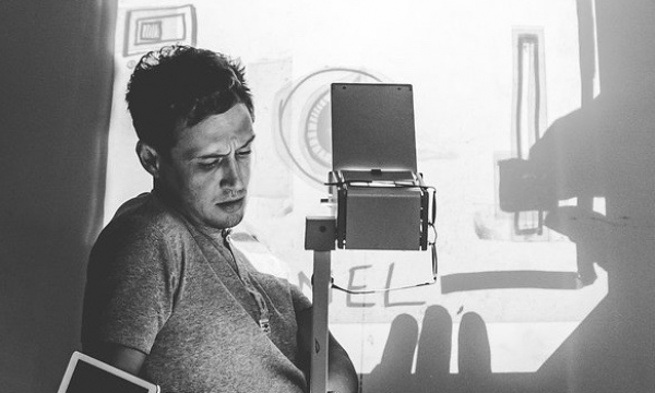 Black and white image of Scott Turnbull sitting behind an overhead projector. An illustration he;s creating is projected onto a screen behind him.