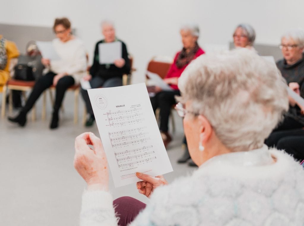 The camera peeks over the shoulder of a woman with short grey hair, she is reading from a piece of sheet music. In the background, other people are seated also reading from sheet music.