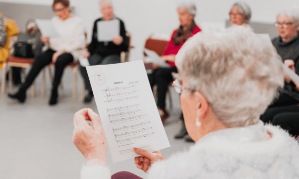 The camera peeks over the shoulder of a woman with short grey hair, she is reading from a piece of sheet music. In the background, other people are seated also reading from sheet music.