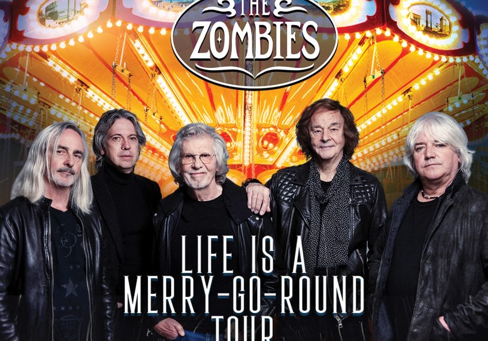 The rock band The Zombies stand in front of a brightly lit merry go round. They all look into the camera. Text in front of them reads "Life Is A Merry Go Round Tour."