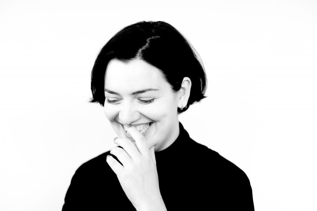 Black and white headshot of Lauren Vevers. Lauren is looking downwards and smiling, and has one hand to her mouth.