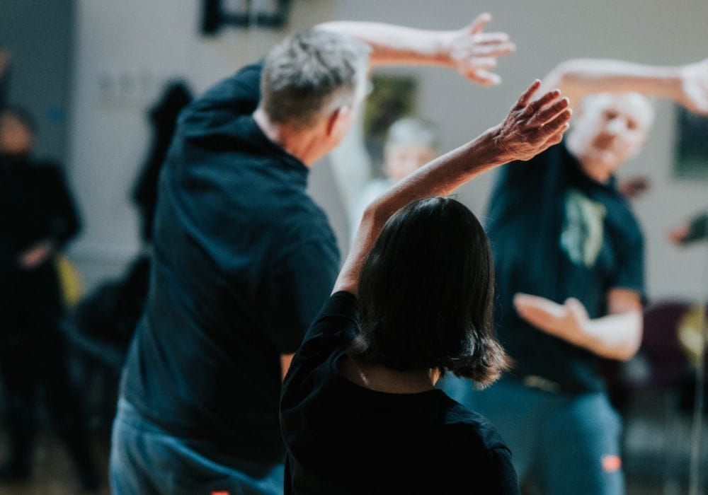 Two people have their back facing the camera, they both have their left arms raised over the heads in a Tai Chi pose.