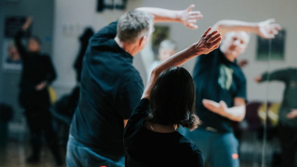 Two people have their back facing the camera, they both have their left arms raised over the heads in a Tai Chi pose.