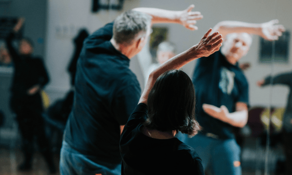 Two people have their back facing the camera, they both have their left arms raised over the heads in a Tai Chi pose.