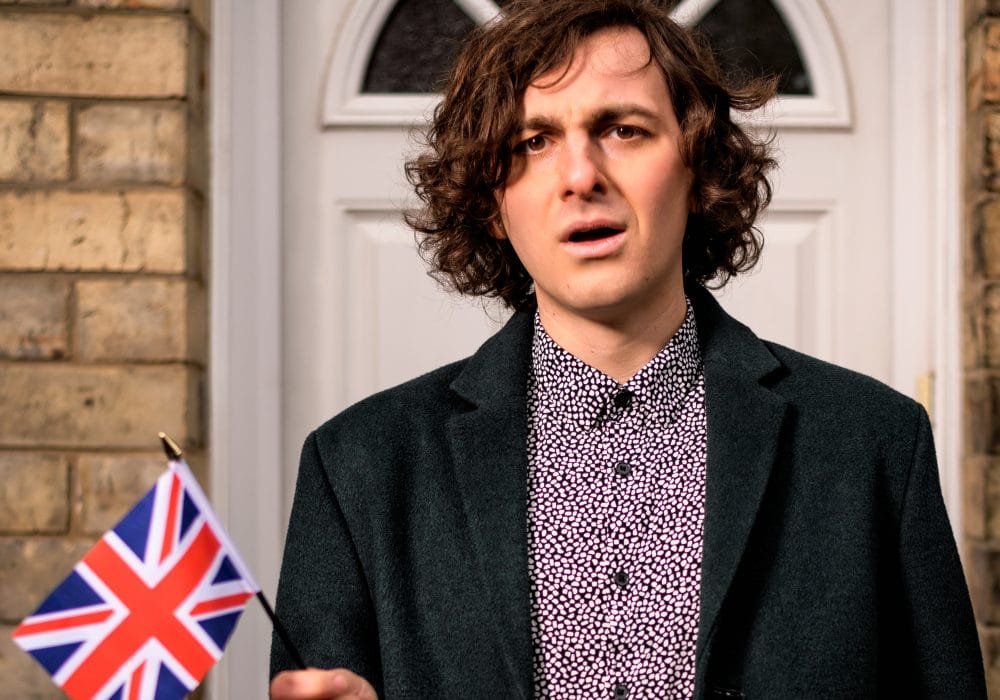 A man with a slightly confused look on his face, waving a small Union Jack, stands in front of a white front door