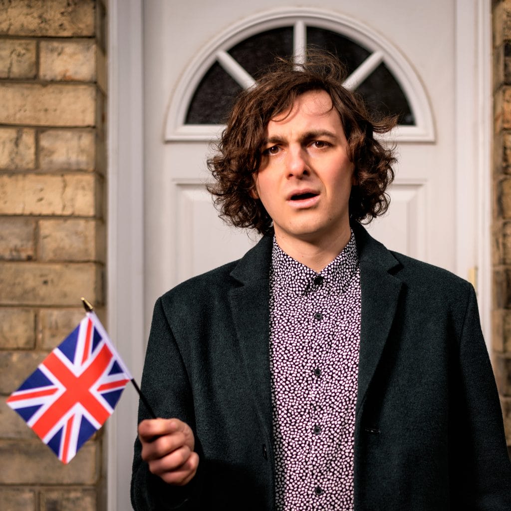 A man with a slightly confused look on his face, waving a small Union Jack, stands in front of a white front door