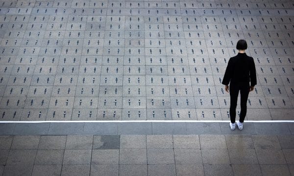 A woman dressed in a black top and trousers, and white trainers, stands with her back to us. In front of her are hundreds of watches laid equally spaced on the floor, two to each grey paving stone.