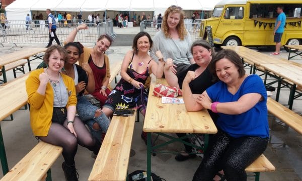 A group of the Tees Women Poets seated at tables and benches and posing for their photo each with one arm in a bodybuilding style pose, to show off their power.