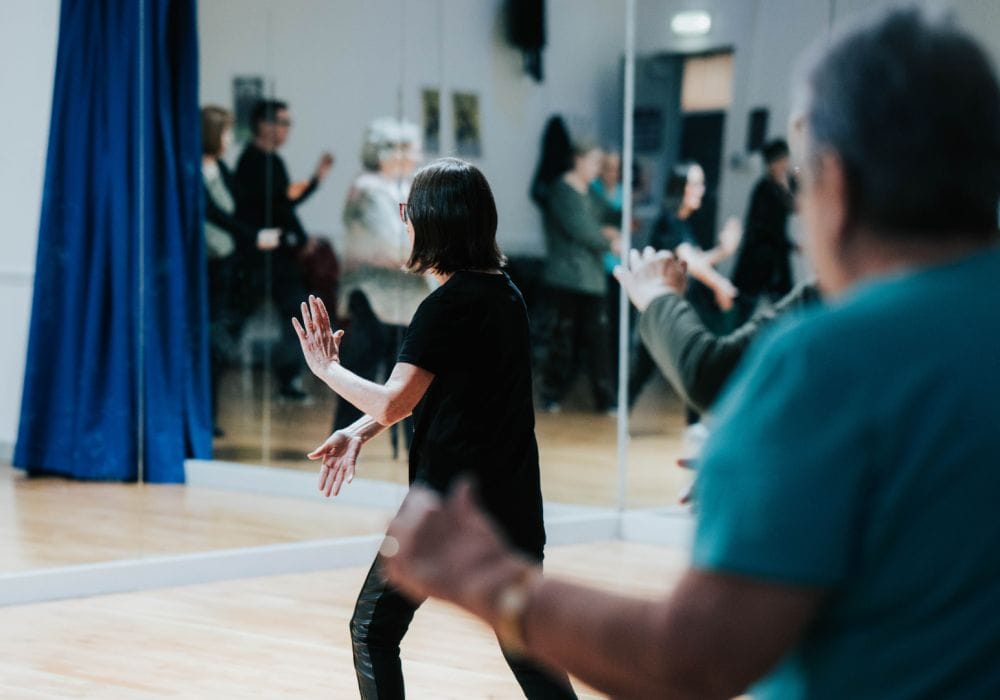 Two people with their backs to the camera, they both have their arms stretched ahead of them in a tai chi pose. The person in the foreground is out of focus, and the person in the background is in focus.