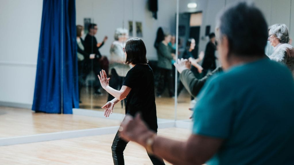 Two people with their backs to the camera, they both have their arms stretched ahead of them in a tai chi pose. The person in the foreground is out of focus, and the person in the background is in focus.