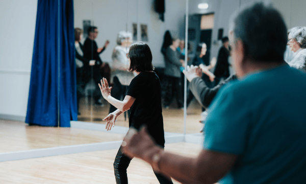 Two people with their backs to the camera, they both have their arms stretched ahead of them in a tai chi pose. The person in the foreground is out of focus, and the person in the background is in focus.