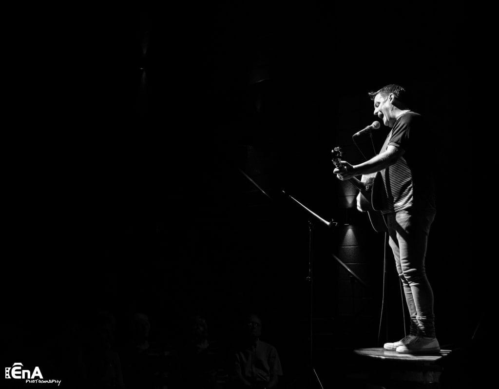 Black and White photo of Mike McGrother standing on a table in a dark room. He has a guitar and is singing into a microphone. by EnA Photography