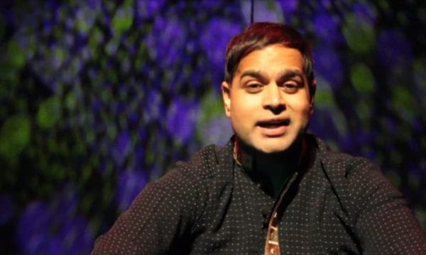 Umar Butt sits in front of a multicoloured background, he is facing the camera and smiling..