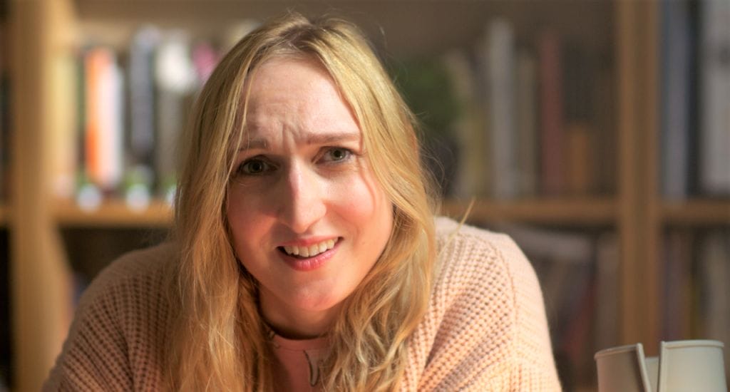 Actor Nicola Chegwin is pictured in a film still from Hen Night, playing the solo lead part of Jessica. Nicola is a young white woman with long blond hair. She is wearing a large oversized pale pink cardigan and pyjamas. She is seated at a desk which is out of shot, but we can see her crutch is leaning against it next to her. She is talking, and looking into the camera quizzically