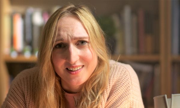 Actor Nicola Chegwin is pictured in a film still from Hen Night, playing the solo lead part of Jessica. Nicola is a young white woman with long blond hair. She is wearing a large oversized pale pink cardigan and pyjamas. She is seated at a desk which is out of shot, but we can see her crutch is leaning against it next to her. She is talking, and looking into the camera quizzically