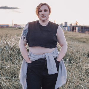 Audrey Cook outside, with Redcar Steelworks behind them. Audrey is wearing a cropped sleeveless black top, black trousers, and has a pale grey jumper tied around their waist