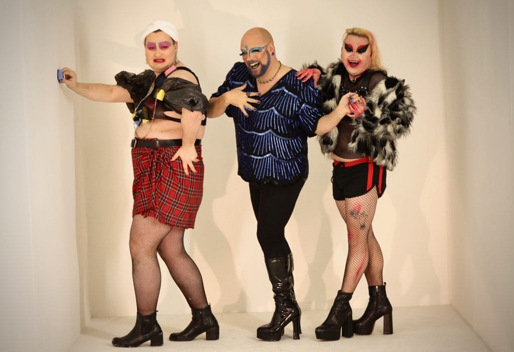 Three drag artists in costume stand in a row and pose for the camera