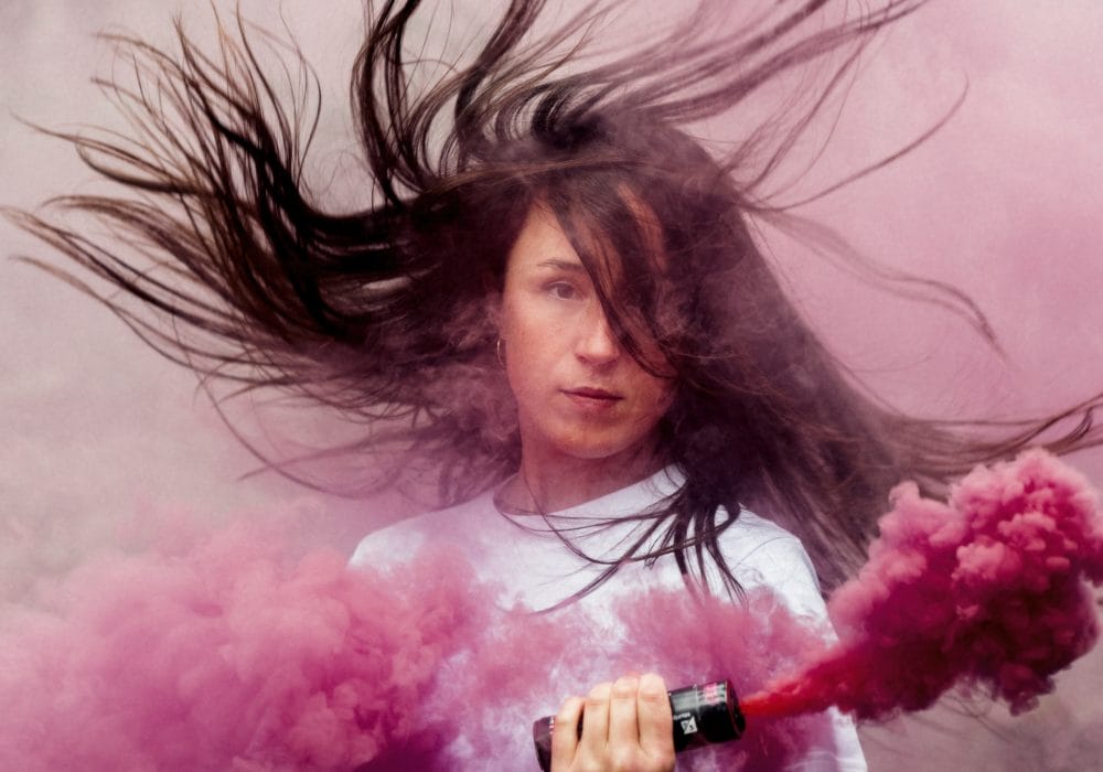 Luca stands facing the camera surrounded by pink smoke, and her long dark hair flying around her head