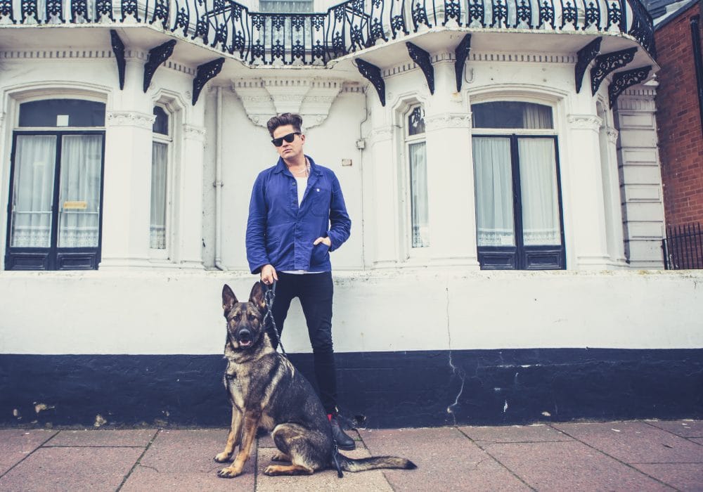 Poet Luke Wright, dressed in dark blue jeans, a white t-shirt, a blue shirt and sunglasses, stands in front of a white building holding a German Shepherd dog on a lead