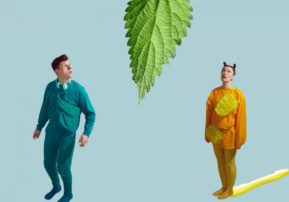 Actors in a green jumpsuit and a yellow jumpsuit dressed up for a children's show as a slug and a caterpillar, looking up at a giant green leaf above them