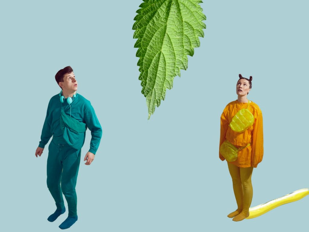 Actors in a green jumpsuit and a yellow jumpsuit dressed up for a children's show as a slug and a caterpillar, looking up at a giant green leaf above them