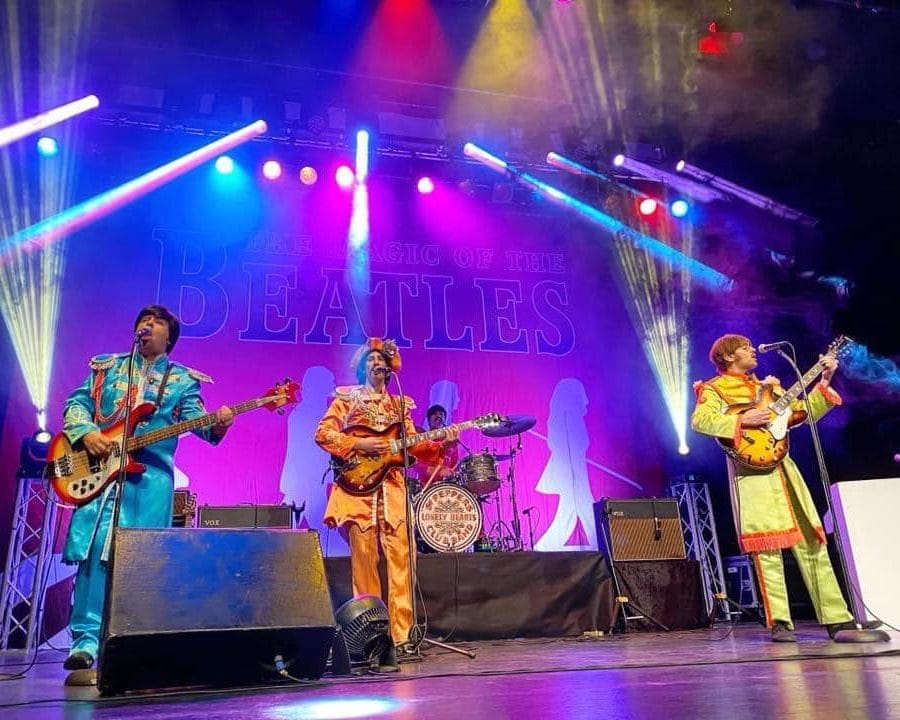 Tribute band Beatlemania stand on a brightly lit stage, they are all holding instruments and wearing bright coloured suits.