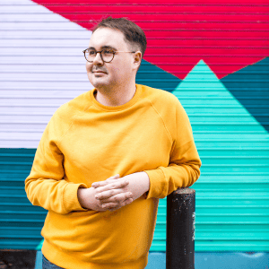 Image of artist Tommy, wearing round glasses and a bright yellow jumper, in front of brightly, multi-coloured painted shutters