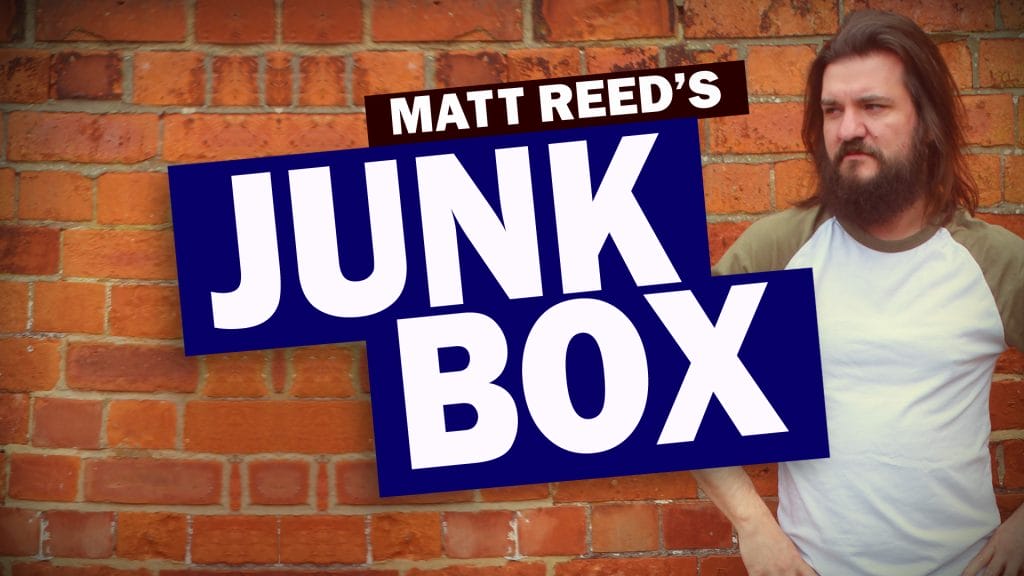 Comedian Matt Reed (shoulder length straight dark hair, beard, wearing a white top with green sleeves) is standing outside against a red brick wall. Text overlaid on the image reads 'MATT REED'S JUNK BOX'