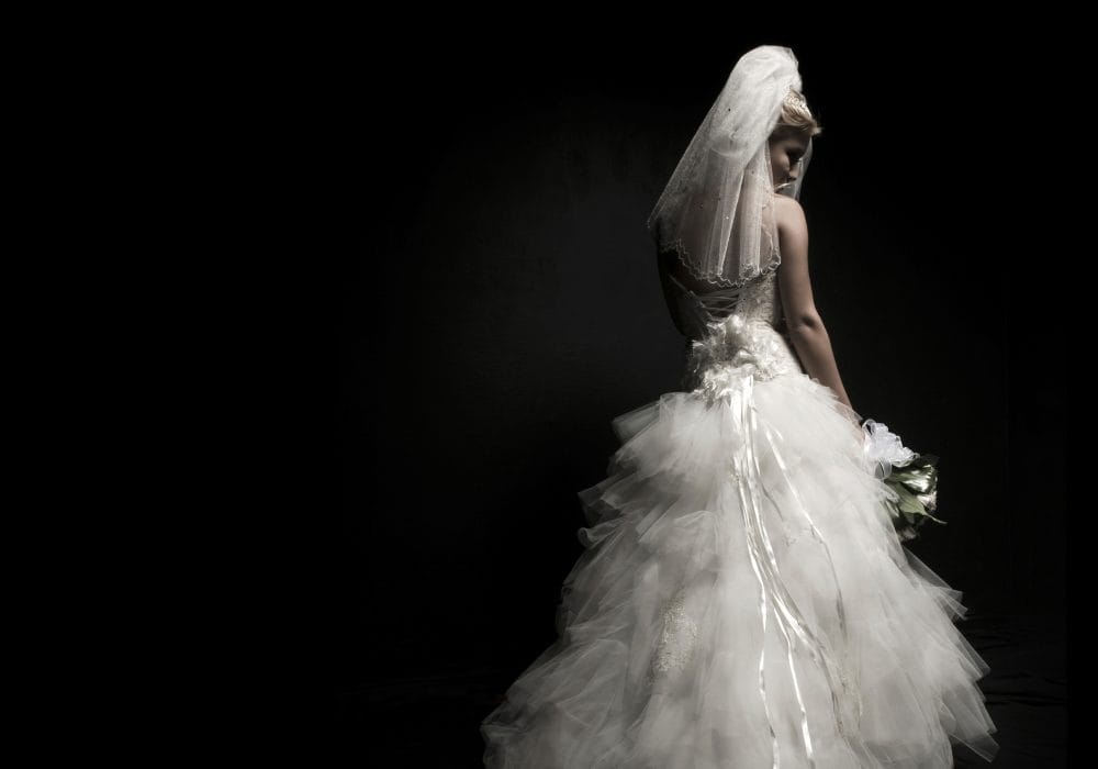 A woman in a white wedding dress and veil, seen from behind, on a black background