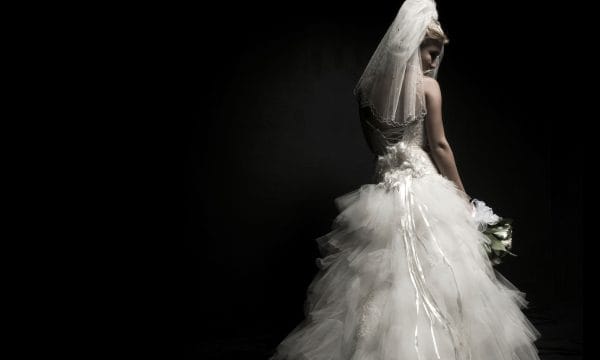 A woman in a white wedding dress and veil, seen from behind, on a black background