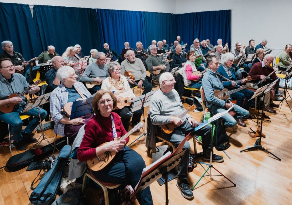 A room packed with people, they are all seated and playing ukuleles.