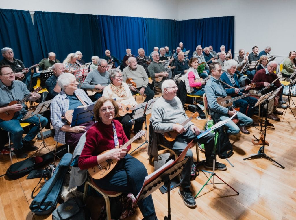 A room packed with people, they are all seated and playing ukuleles.