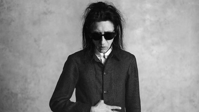 Black and white image of John Cooper Clarke. He has long dark hair, and is wearing sunglasses and a dark jacket buttoned almost to the top. He is looking down towards the floor.