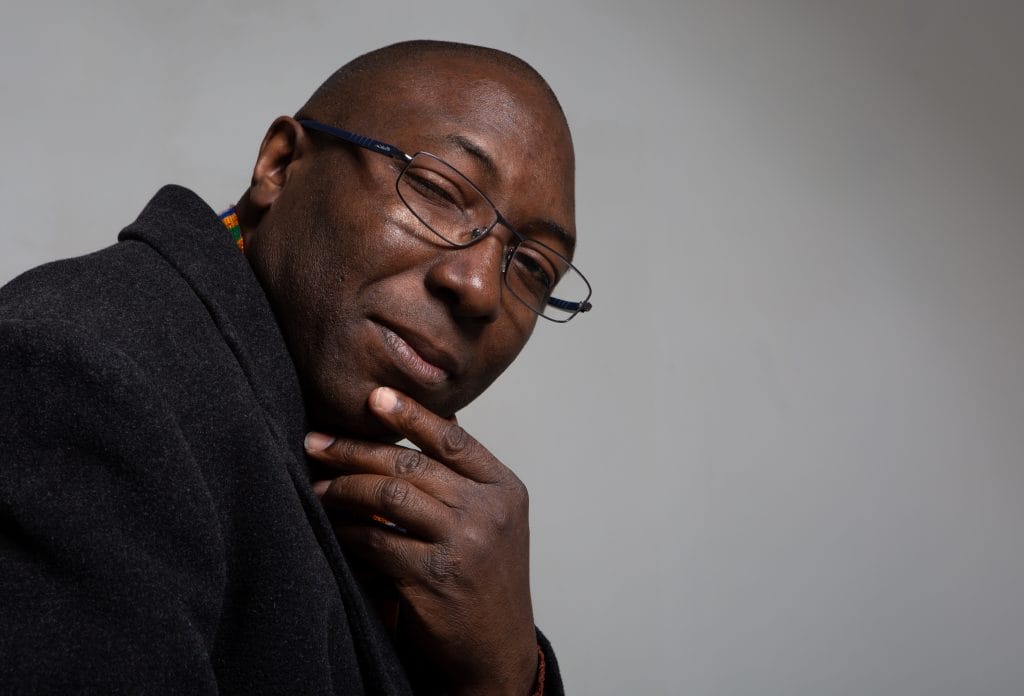 A headshot of artist Dominic JP Nelson-Ashley, he is looking thoughtfully into the camera lens, with one hand on his chin.