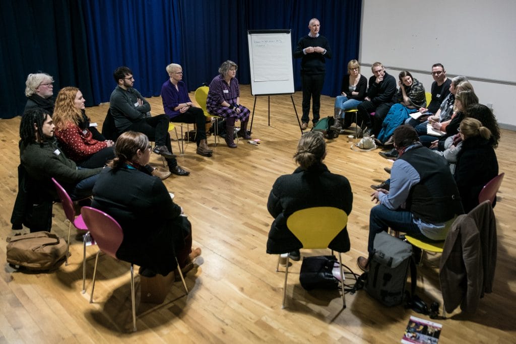 A group of 17 people sit in a circle (on chairs, some pink, some yellow) around a speaker standing next to a flip chart with notes on it. The room they're in - ARC's dance studio - has plain white walls, a wooden floor, and dark blue curtains down one side.