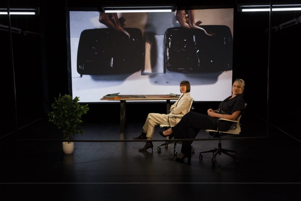 Two female performers sit in office style chairs in front of a table on a black box style theatre set. They are both on the same side of the set and are in the same pose, looking side on towards the audience. There is a large pot plant on the front left of the set, and a projection screen in the background.
