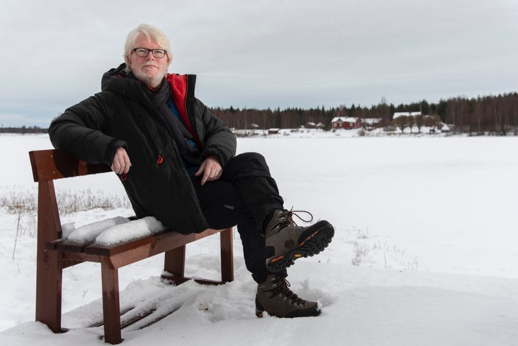 Artist Aidan Moseby (white hair, white beard, black framed rectangular glasses) sits on a small wooden snow-covered bench in a snowy field against a plain grey sly, He is wearing chunky walking boots and warm winter clothing (trousers and jacket).